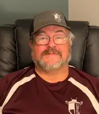 Portrait of a person wearing glasses, a dark baseball cap, and a maroon athletic shirt, seated in a black recliner chair and facing the camera indoors.