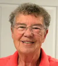 Headshot of a person with short curly hair and glasses wearing a coral-colored collared shirt, photographed indoors against a light-colored background.