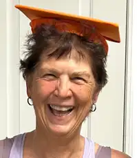 Headshot of a person with short dark hair wearing a light-colored sleeveless top and an orange graduation cap, standing in front of a light-colored paneled background.