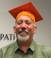 Person wearing an orange graduation cap and matching tassel, standing indoors in front of an office wall.